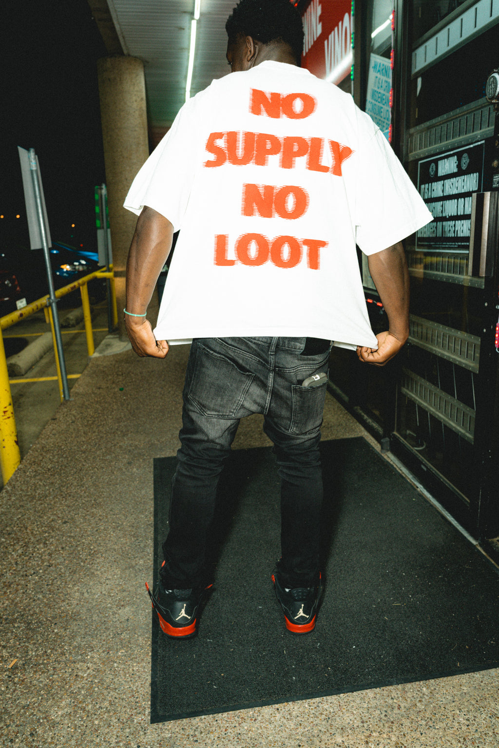 oversized white streetwear tee "no supply no loot"  wearing a white t-shirt with 'NO SUPPLY NO LOOT' text, standing on a sidewalk at night.