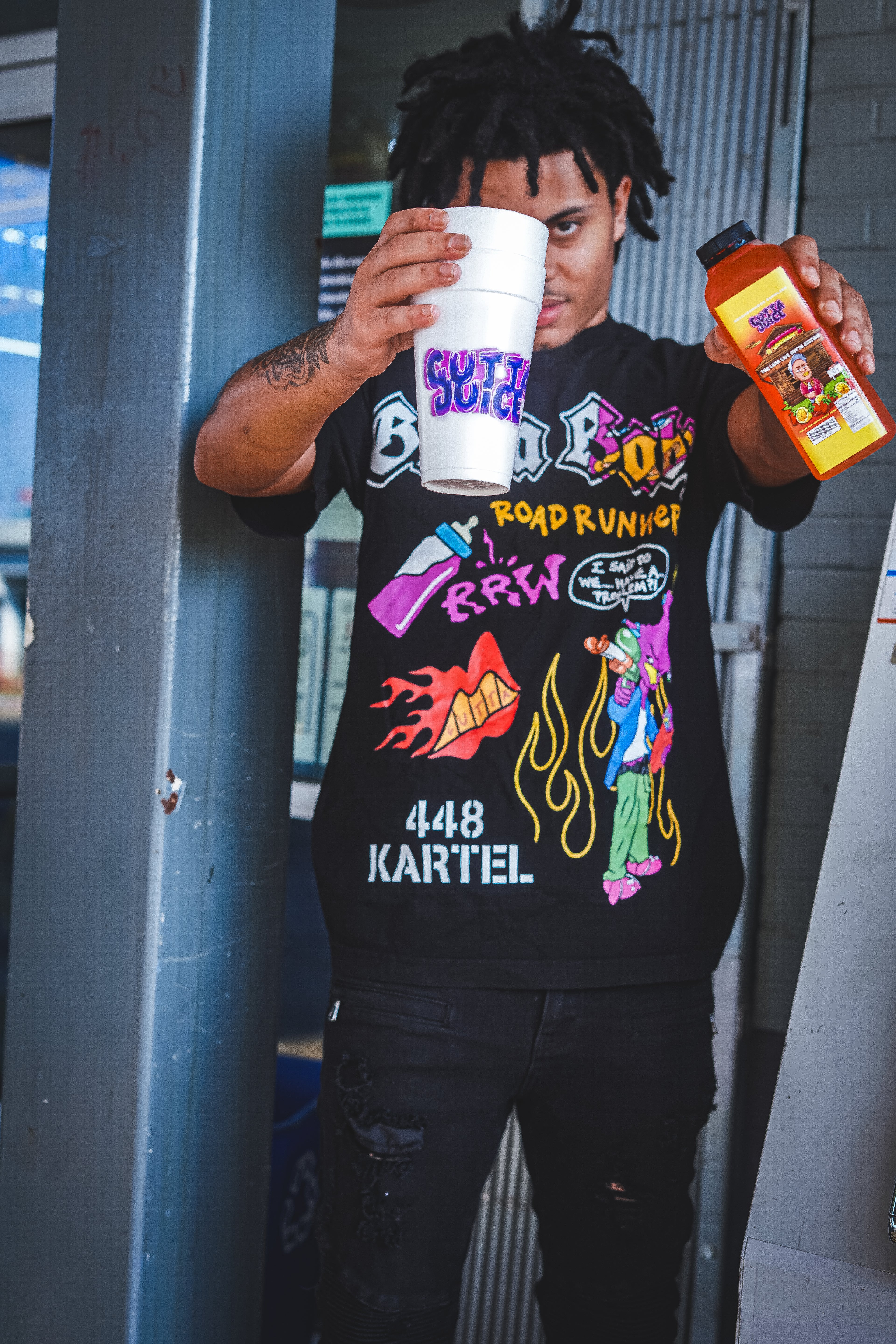 A person holding a white styrofoam cup with 'GUTTA JUICE' text and colorful graphics on a black T-shirt with neighborhood supplier gutta juice lemonade and gutta juice styrofoam double cups